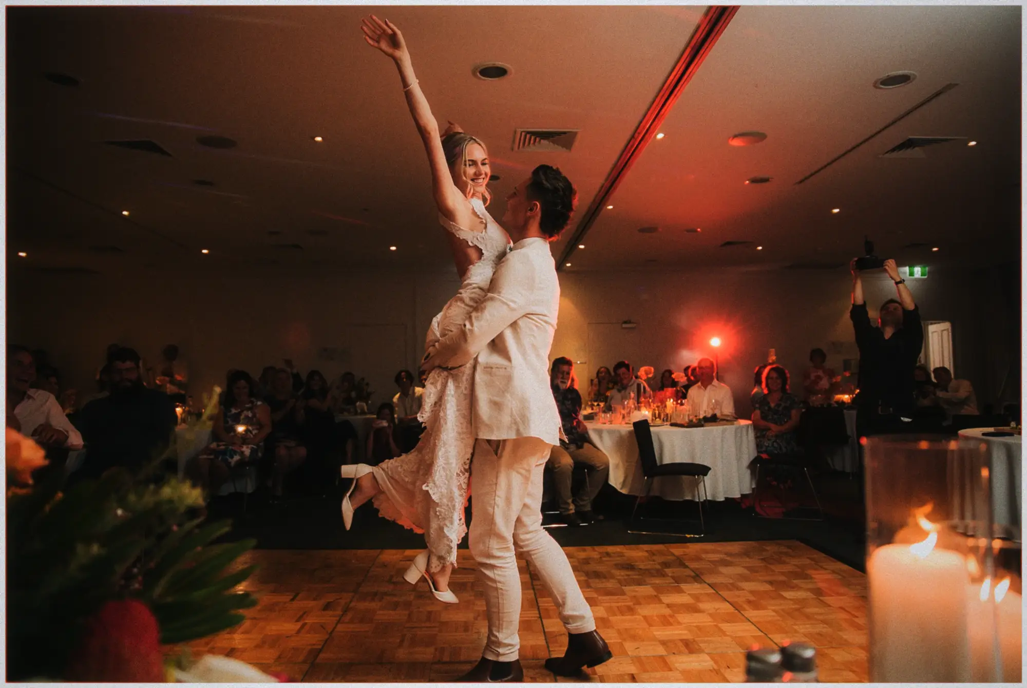 Justin lifts Erin overhead mid-dance, her arm stretched to the ceiling as guests cheer.