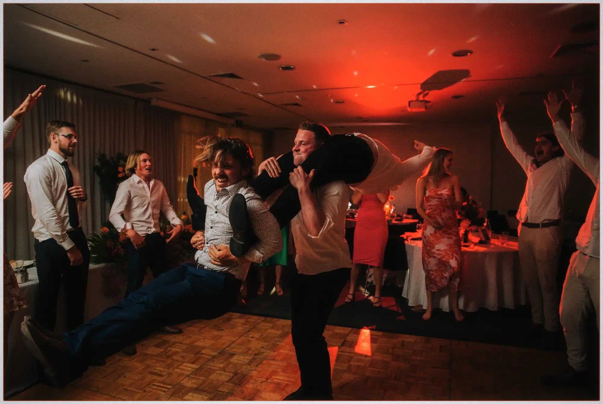 Groomsmen piggyback roughhouse across the floor in red party light as guests cheer.