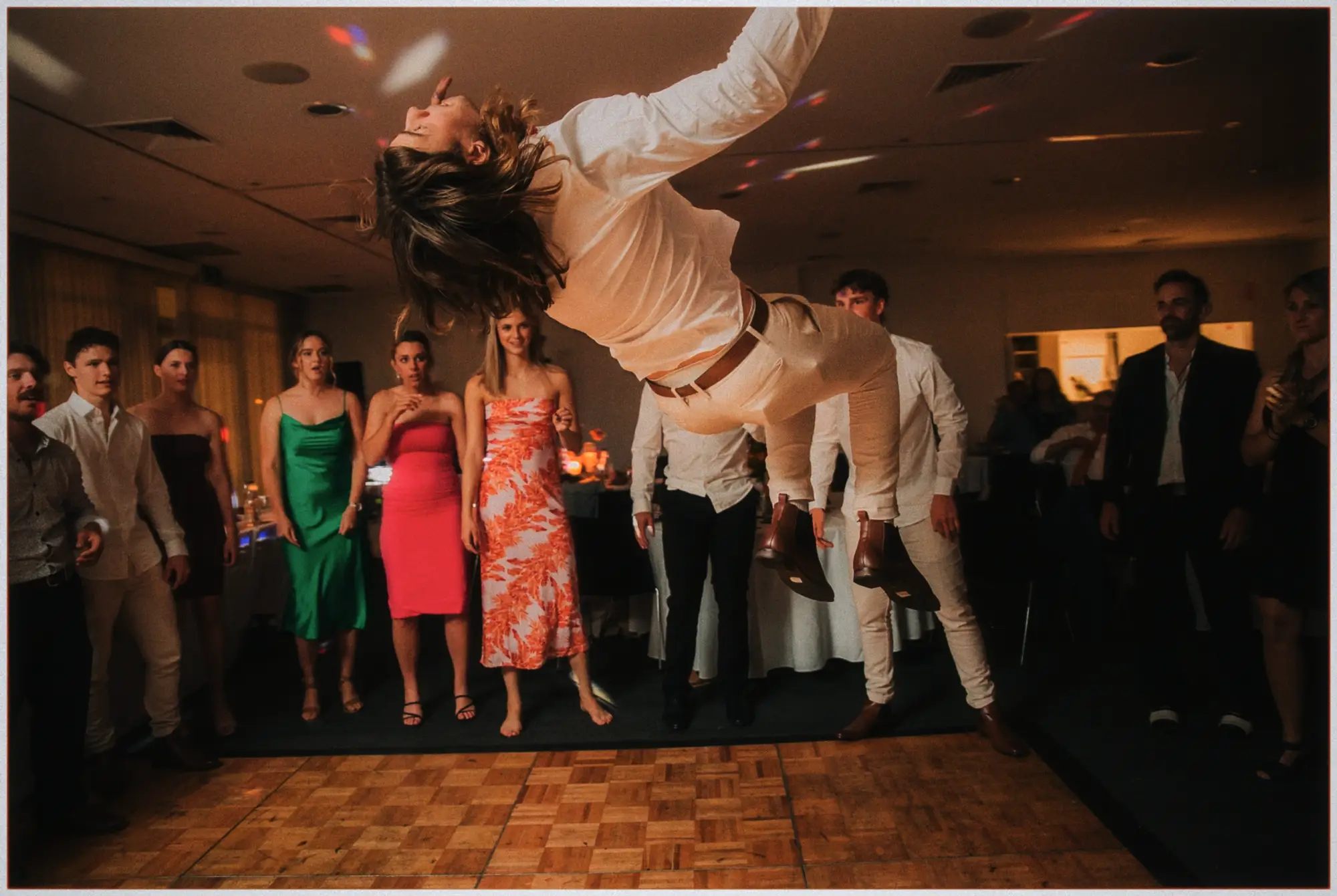 A guest backflips across the dance floor while partygoers watch in warm evening light.