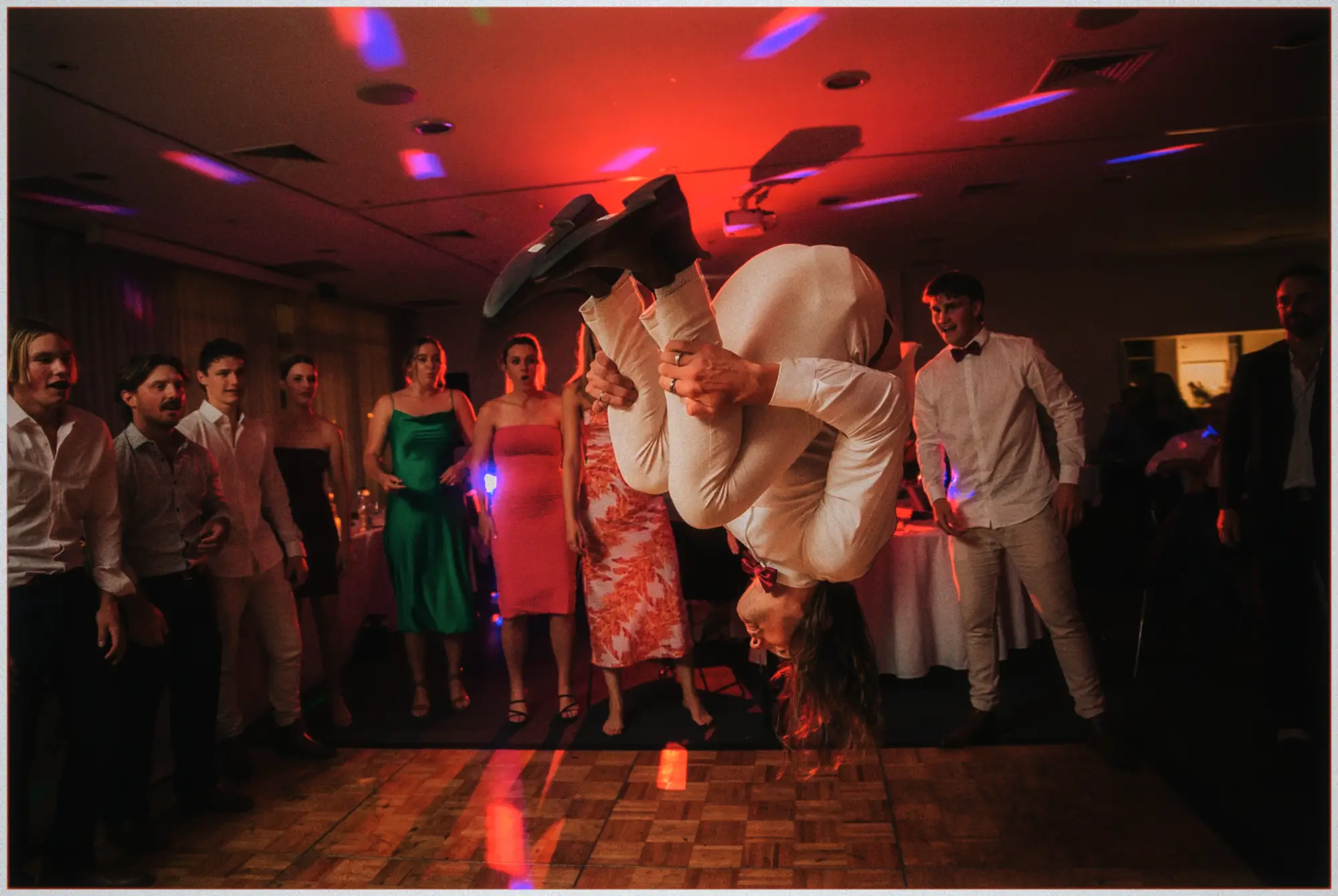 Groom in a white suit mid backflip on the dance floor as stunned guests watch under red lights