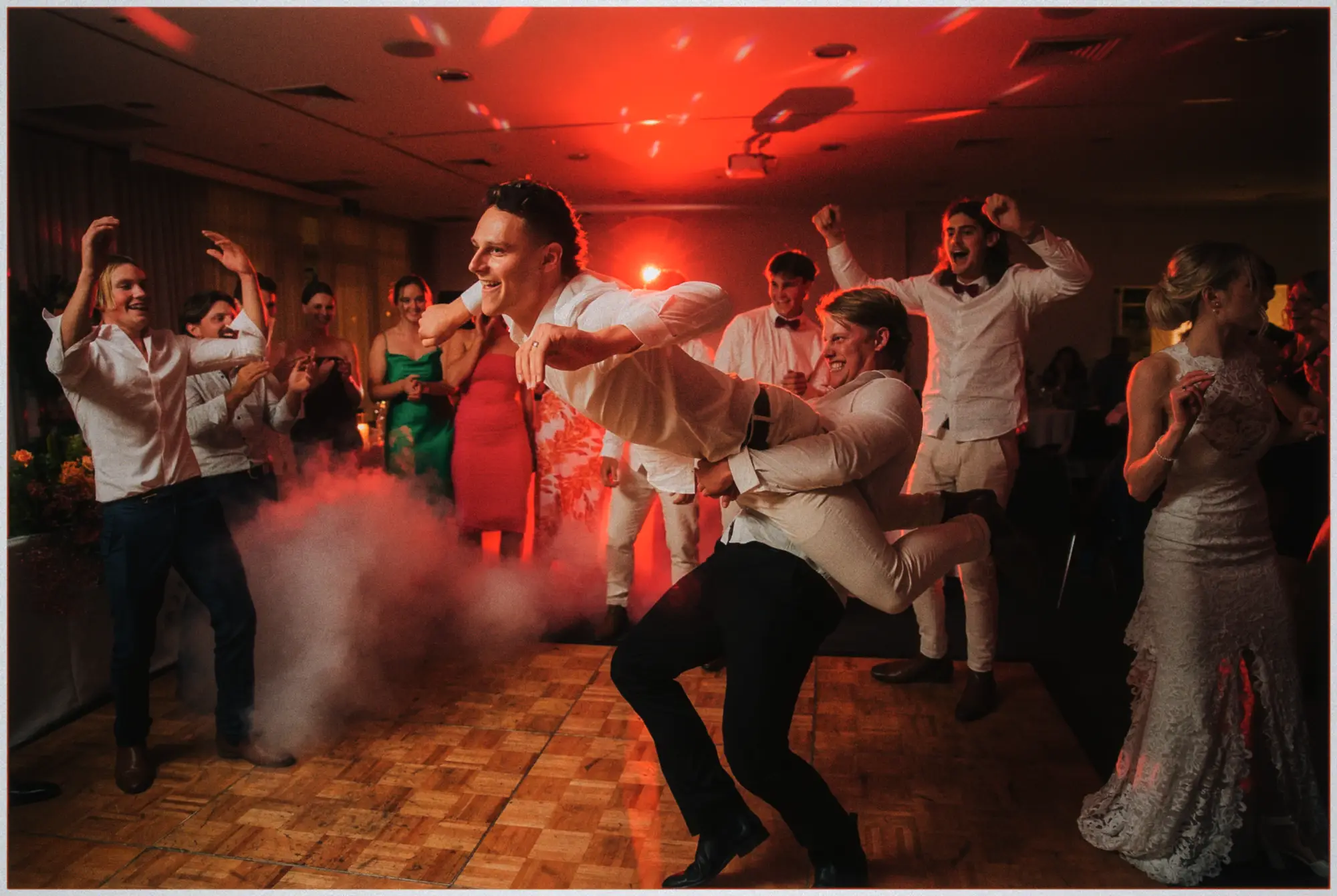 Friends dipping the groom backwards through smoke as guests cheer and the bride laughs in her gown